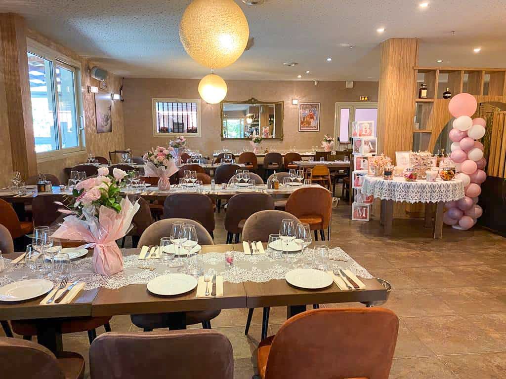Une salle à manger dressée pour une fête avec des centres de table floraux roses, des chemins de table en dentelle blanche et une arche de ballons roses.