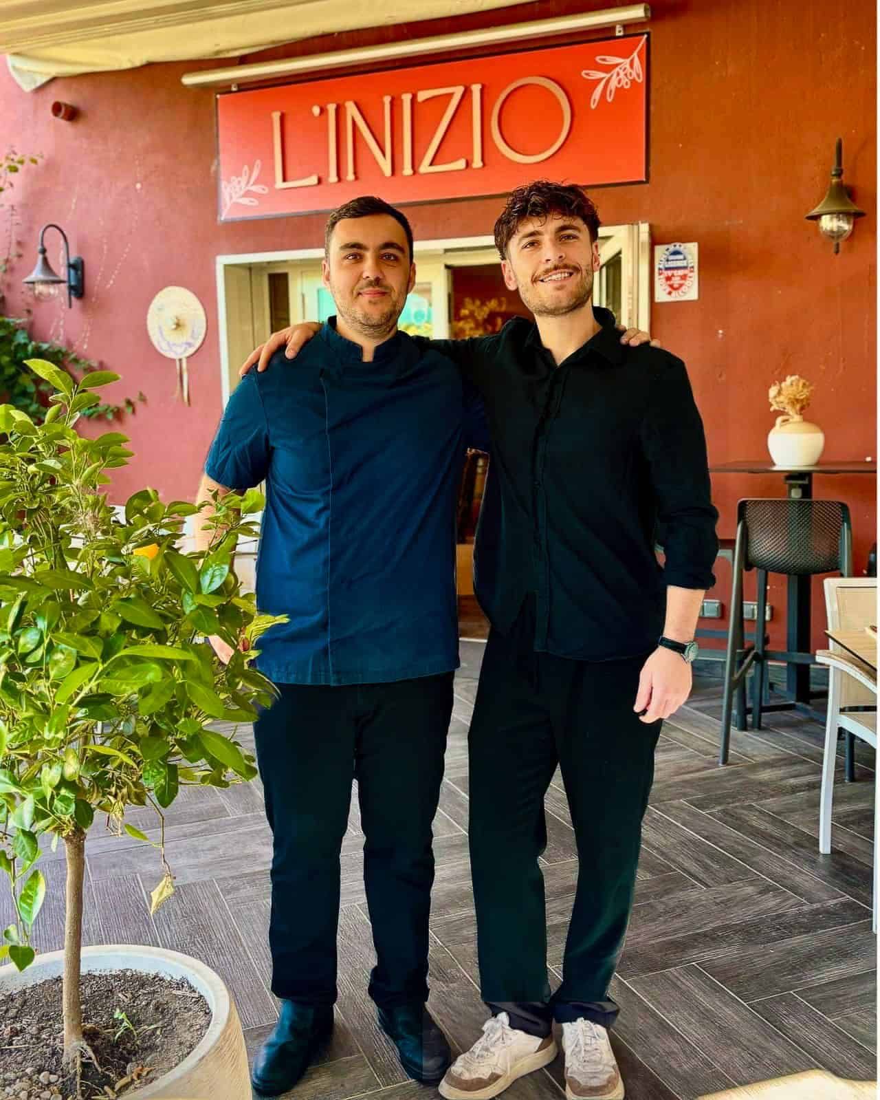 Deux hommes souriants, bras dessus bras dessous, devant un restaurant appelé L'INIZIO, avec un citronnier au premier plan.
