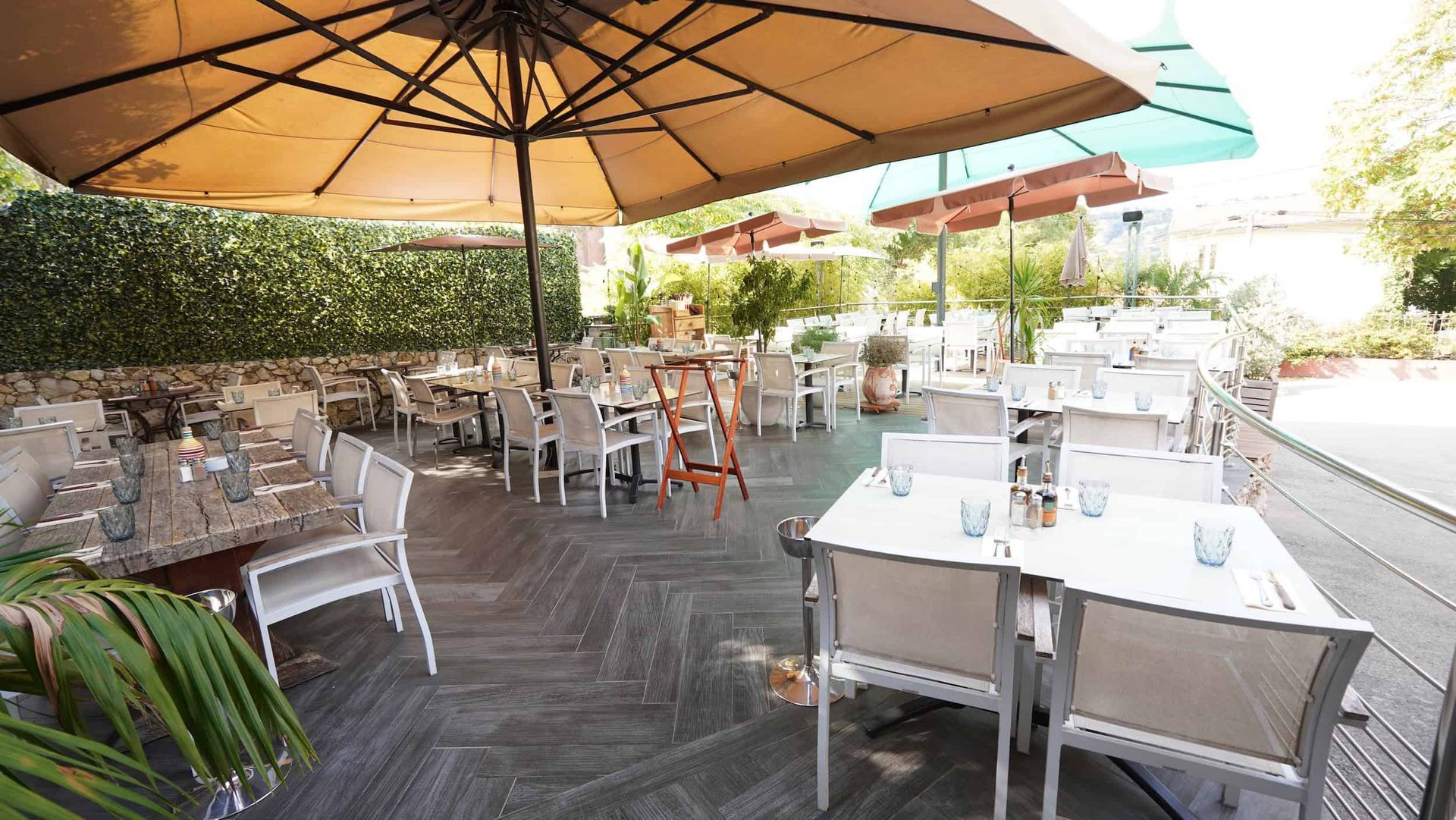 Une terrasse de restaurant en plein air avec des chaises blanches, des tables à manger et de grands parasols au-dessus d'une terrasse en bois sombre.