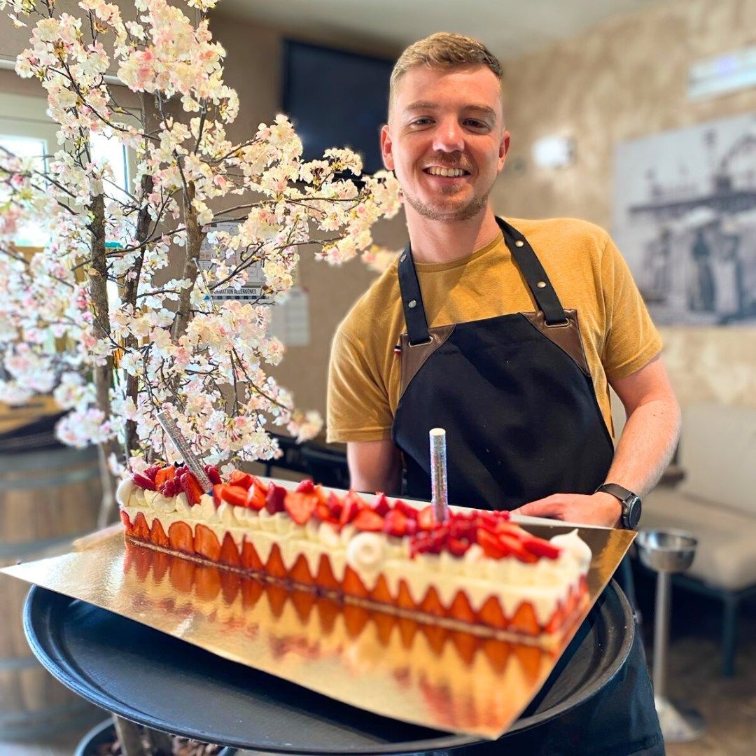 Un serveur souriant, vêtu d'une chemise jaune et d'un tablier noir, présente un long gâteau rectangulaire aux fraises sur un plateau doré.