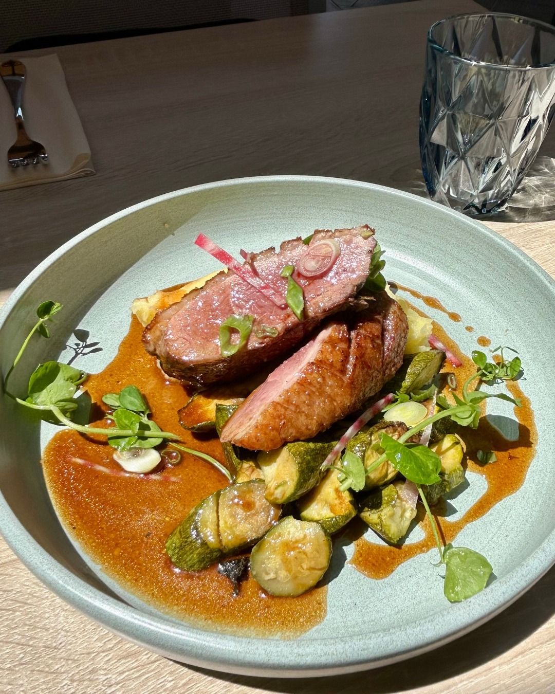 Un plat de magret de canard poêlé, servi avec des courgettes rôties et une riche sauce brune dans un bol vert clair, sur une table en bois.
