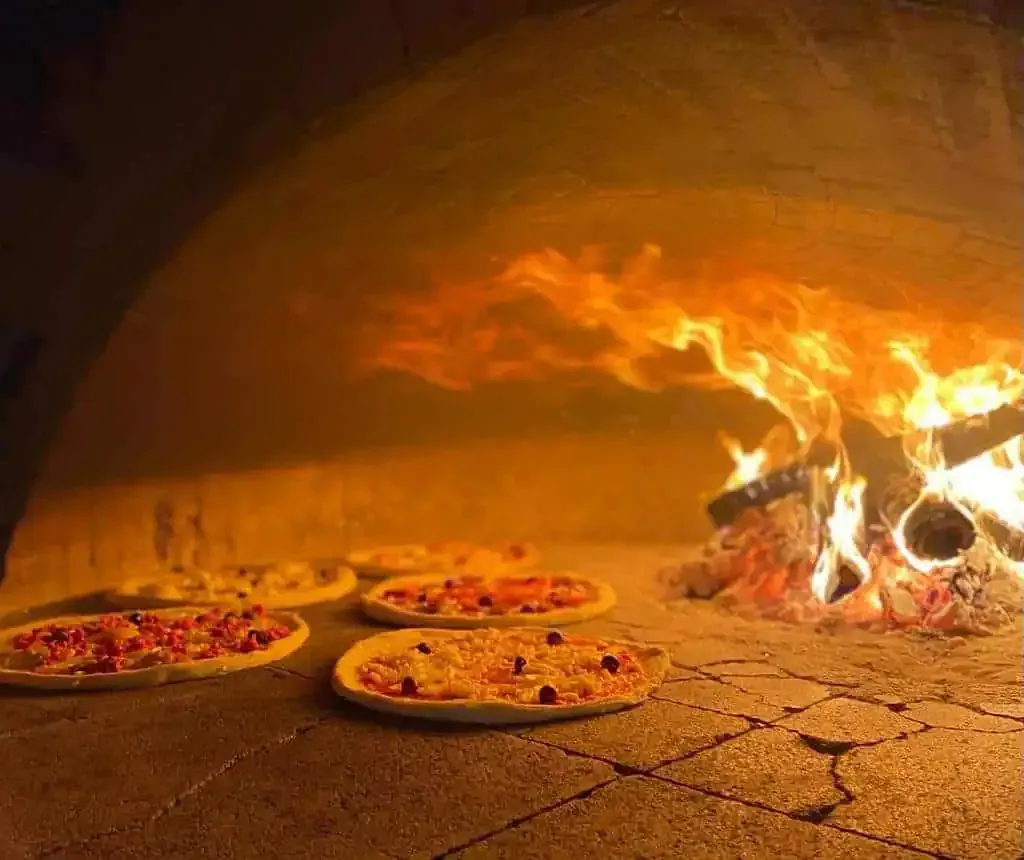 Plusieurs pizzas cuisent dans un four à bois en pierre, avec un feu encore actif à droite.