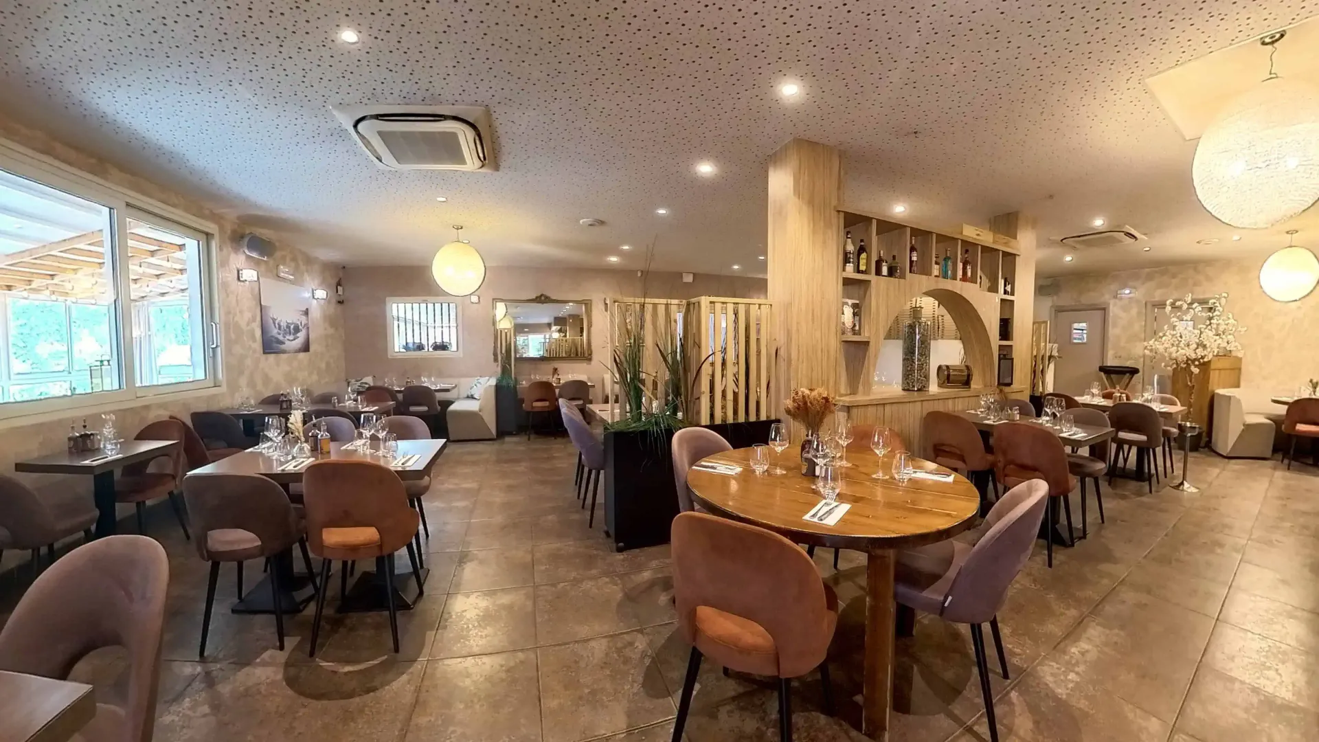 Une salle à manger de restaurant bien éclairée, avec de nombreuses tables rondes et carrées en bois et des chaises rembourrées sur un sol carrelé.