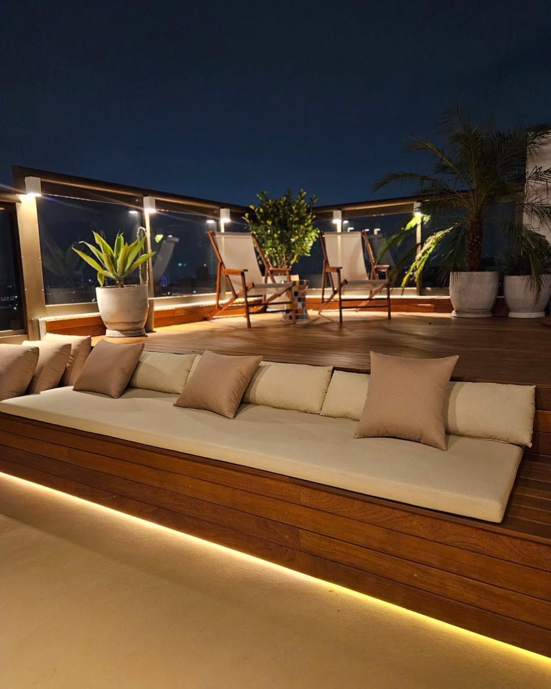 Um terraço moderno na cobertura à noite, com dois longos banco de madeira, almofadas cor creme e iluminação embutida.