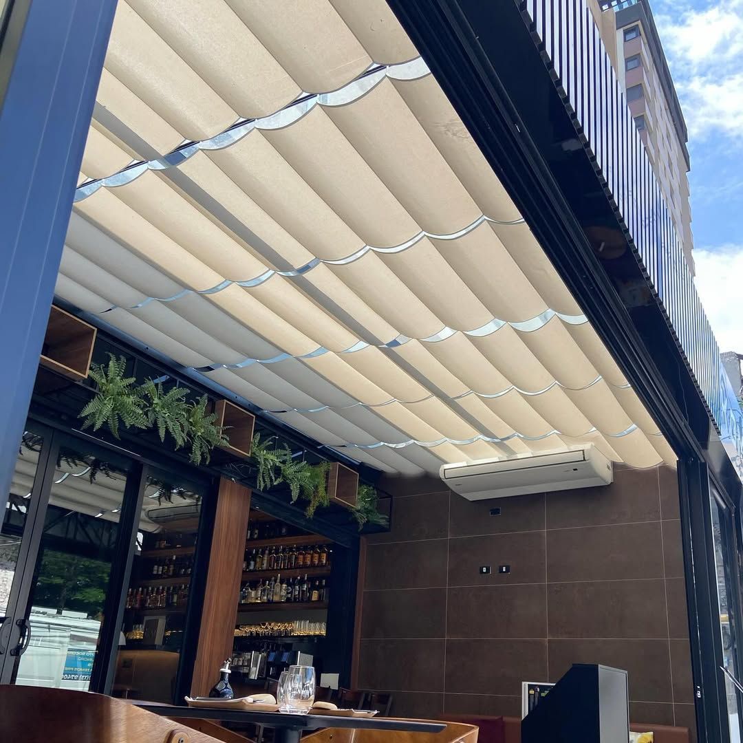 Um pátio externo de restaurante com cobertura retrátil bege, vegetação suspensa e uma unidade de ar condicionado instalada.
