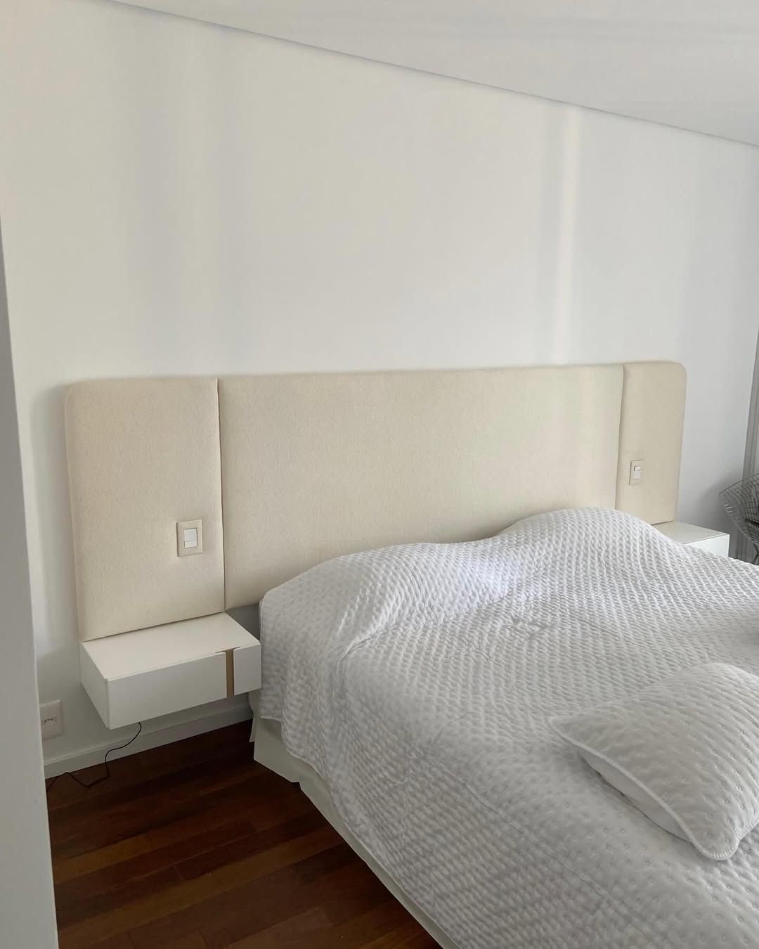 Cabeceira estofada em tom creme com mesas de cabeceira suspensas integradas e interruptores de luz fixados na parede.