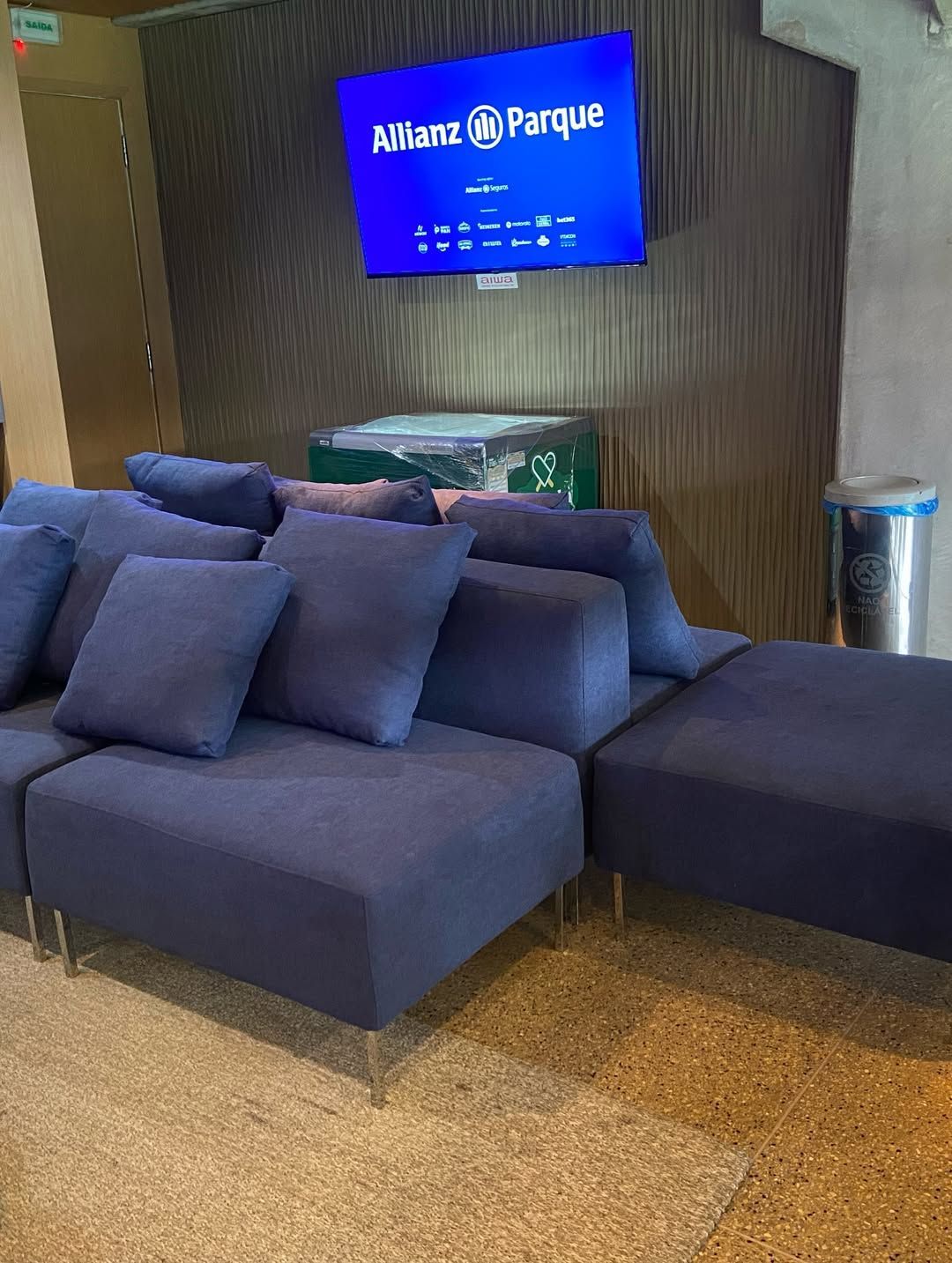 Sofá modular azul localizado em uma área de estar abaixo de uma TV fixada na parede, exibindo a logo do Allianz Parque.