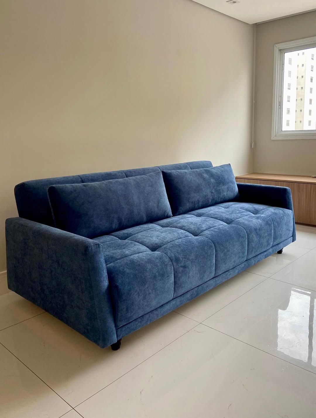 Um sofá azul com capitonê está sobre um piso de azulejos brancos, encostado em uma parede lisa ao lado de uma janela.
