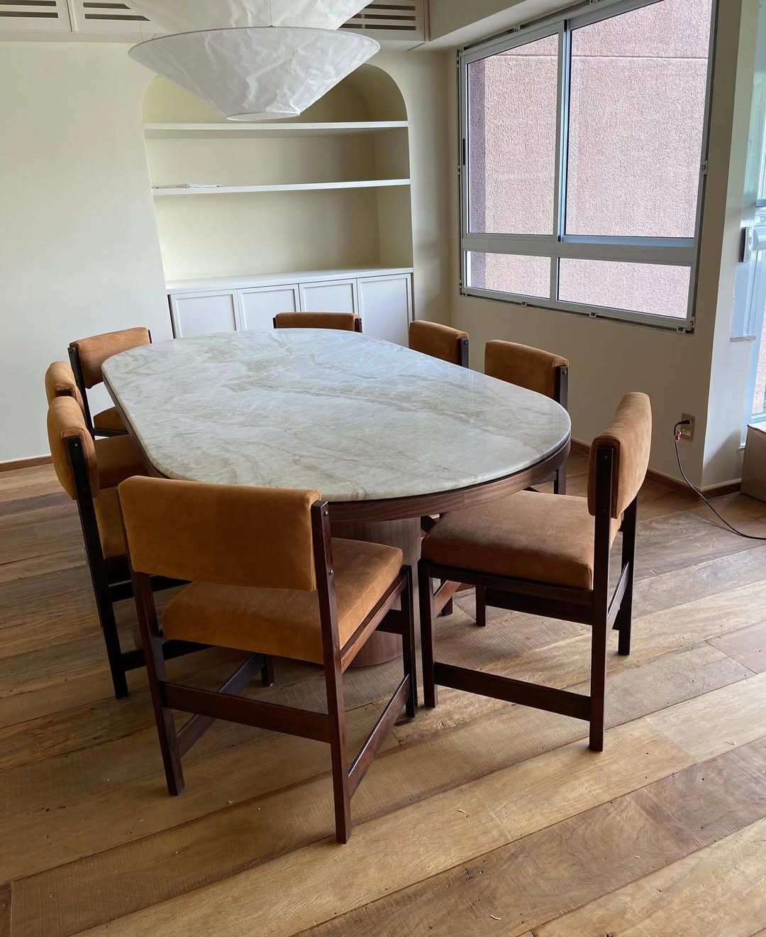 Conjunto de jantar com uma mesa oval clara e seis cadeiras estofadas em marrom sobre um piso de madeira perto de uma janela.