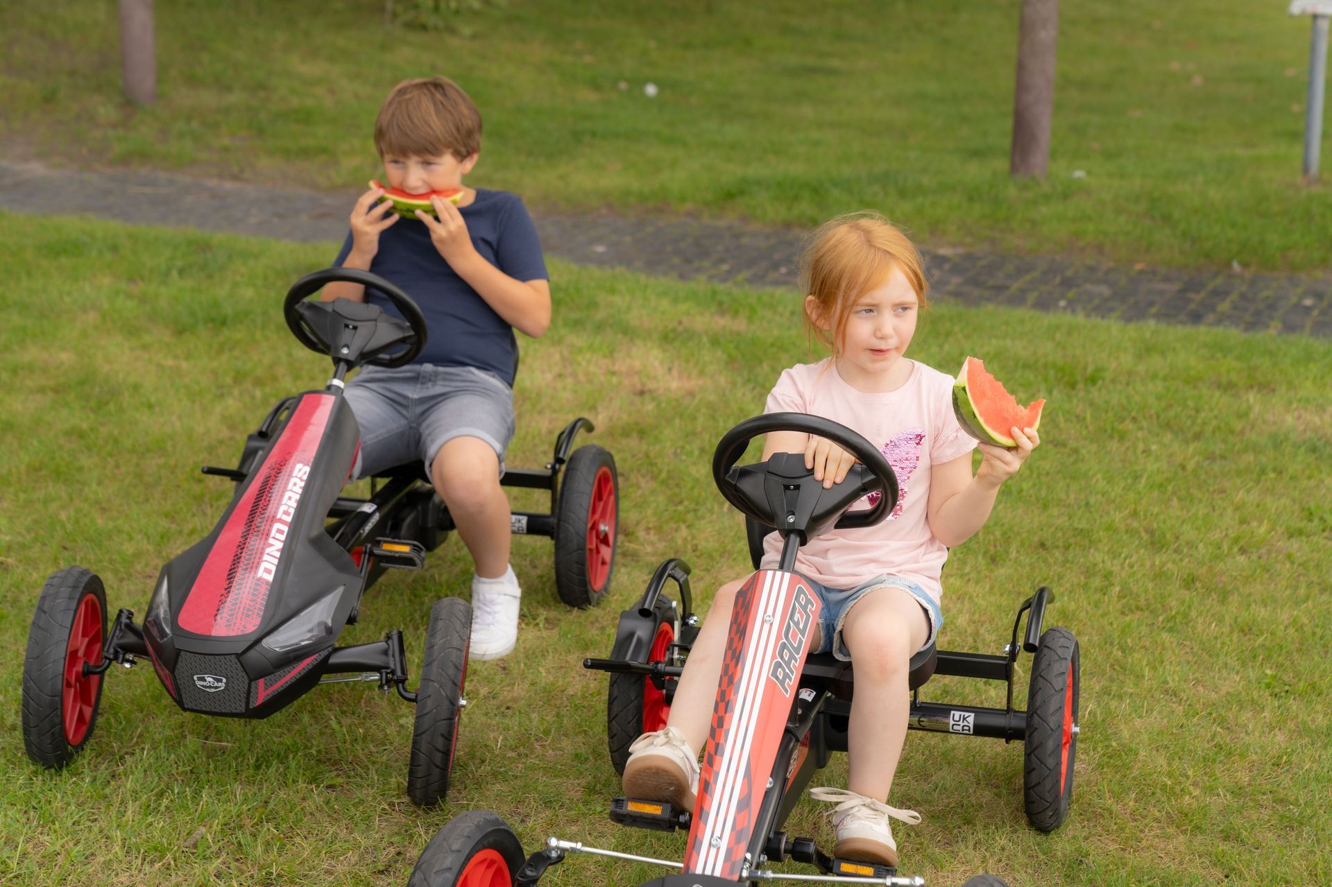 Zwei Kinder fahren mit Pedal-Gokarts auf einer Wiese und essen dabei Wassermelonenscheiben.