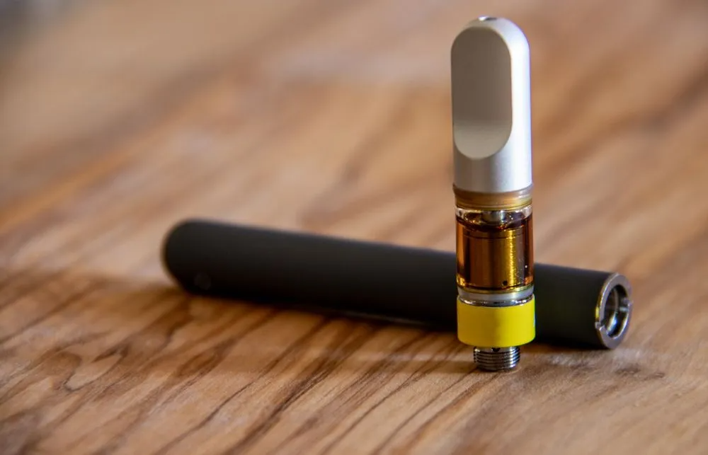 A vape pen with a cartridge on a wooden surface. The cartridge contains amber liquid.