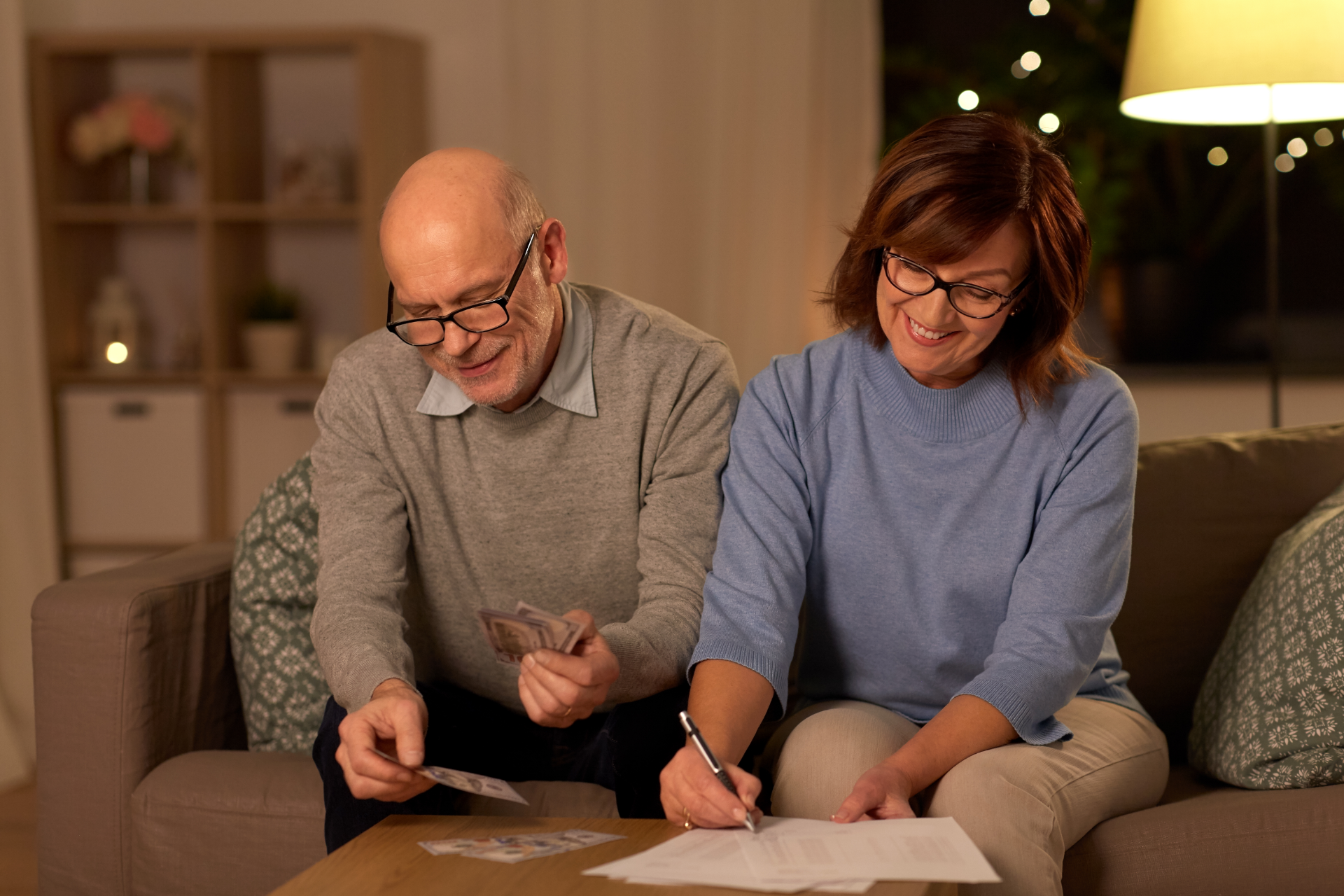 Two people writing on papers at a table in a cozy living room, with a warm lamp and holiday lights in the background