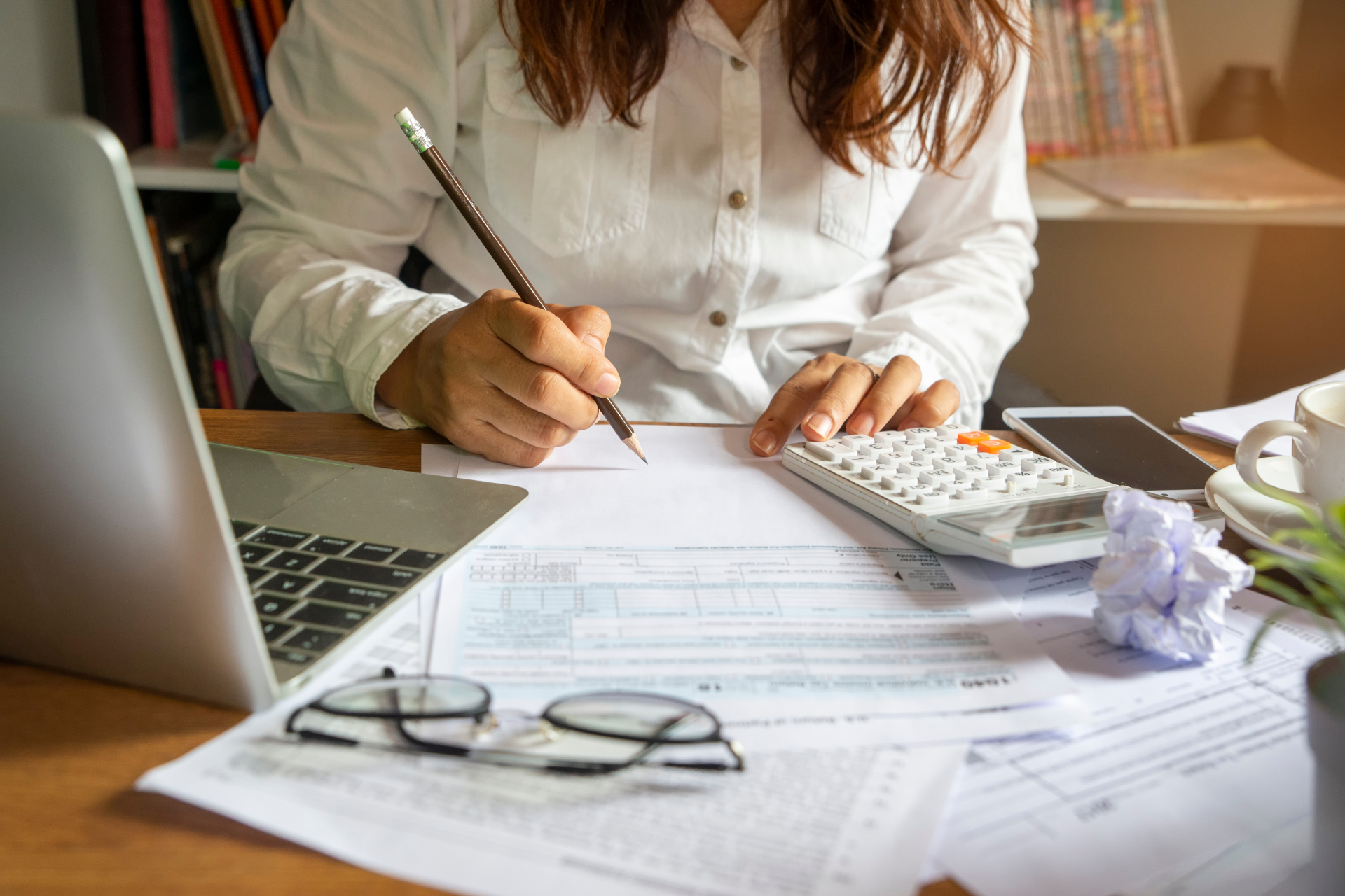 Person writing on paperwork at a desk with laptop, calculator, glasses, and crumpled note