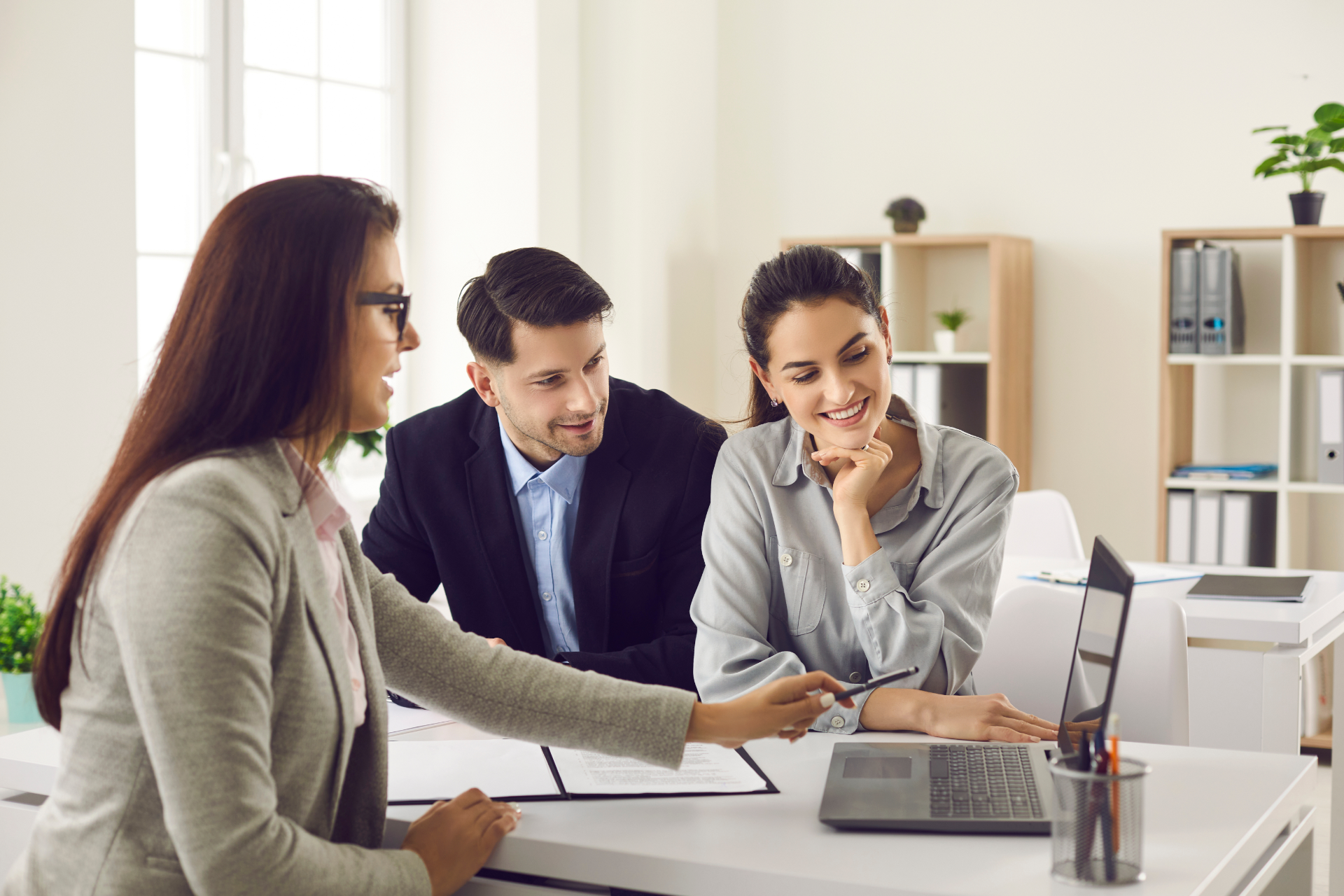 Three coworkers reviewing a laptop at a desk in a bright office, one pointing at the screen.