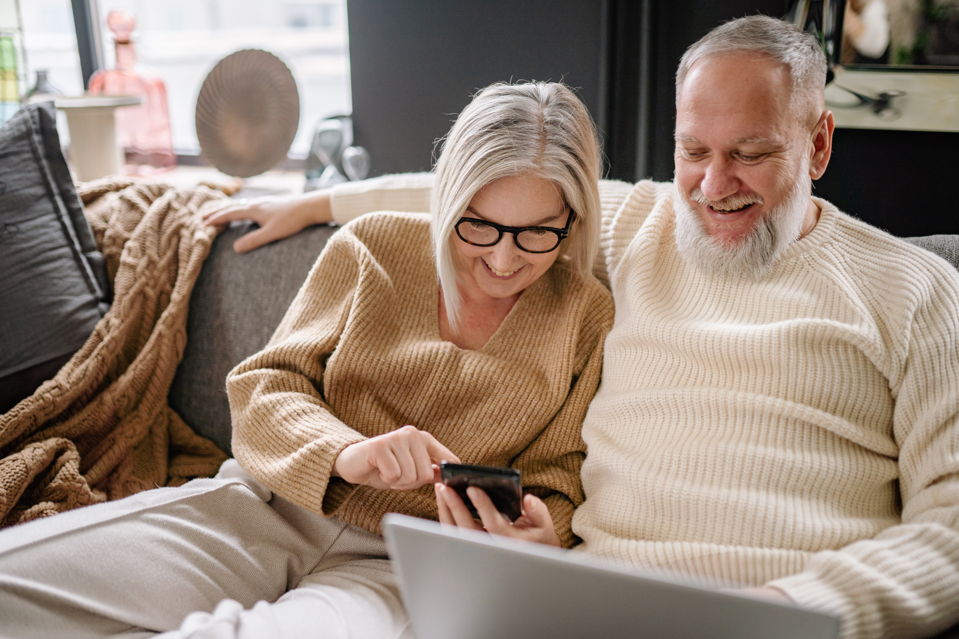 Two people in cream sweaters smile while looking at a tablet on a couch with a laptop nearby