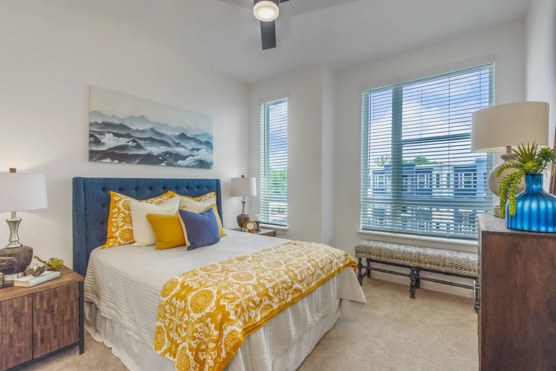 Bedroom with a blue tufted headboard, white bedding, yellow pillows, two nightstands, and windows with blinds.