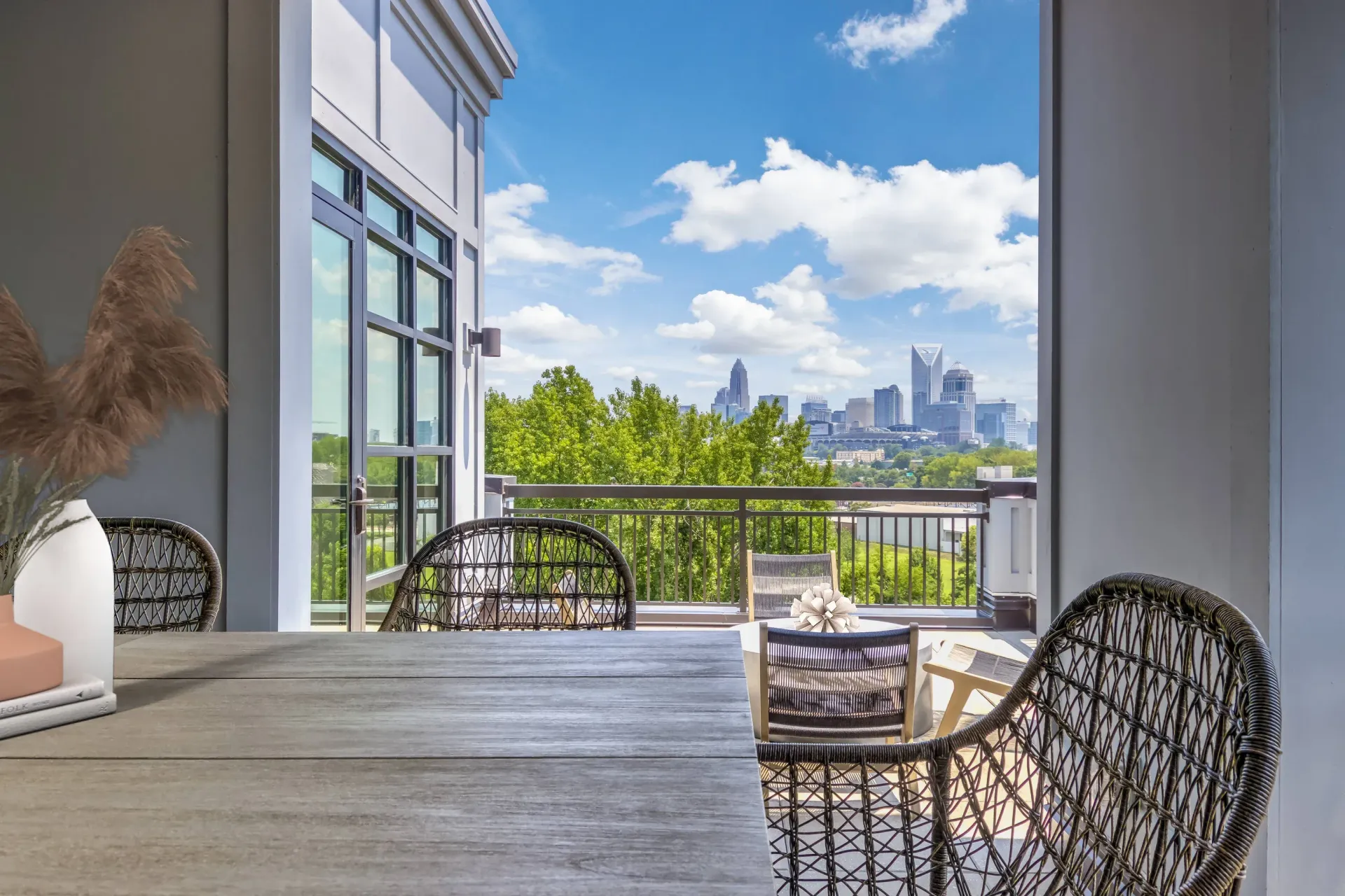 Balcony with a wooden table, woven chairs, and a railing, overlooking a city skyline.