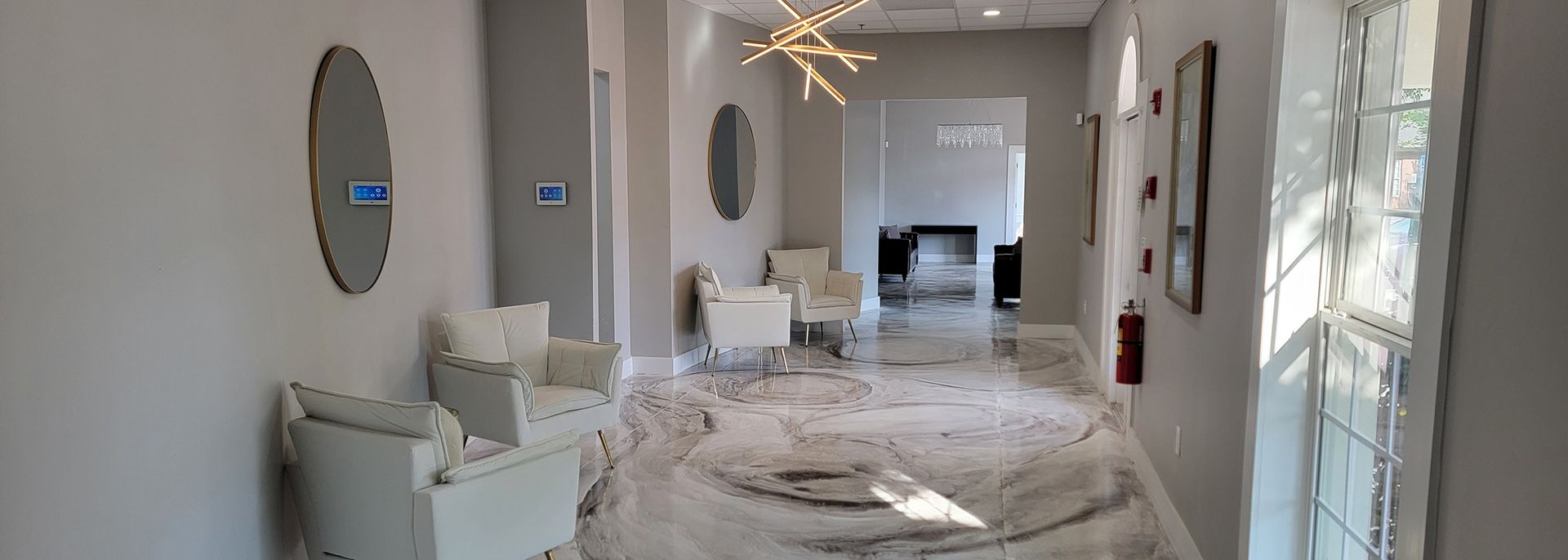 Modern hallway with white chairs, round mirror, chandelier, and a rug leading to a bright room.
