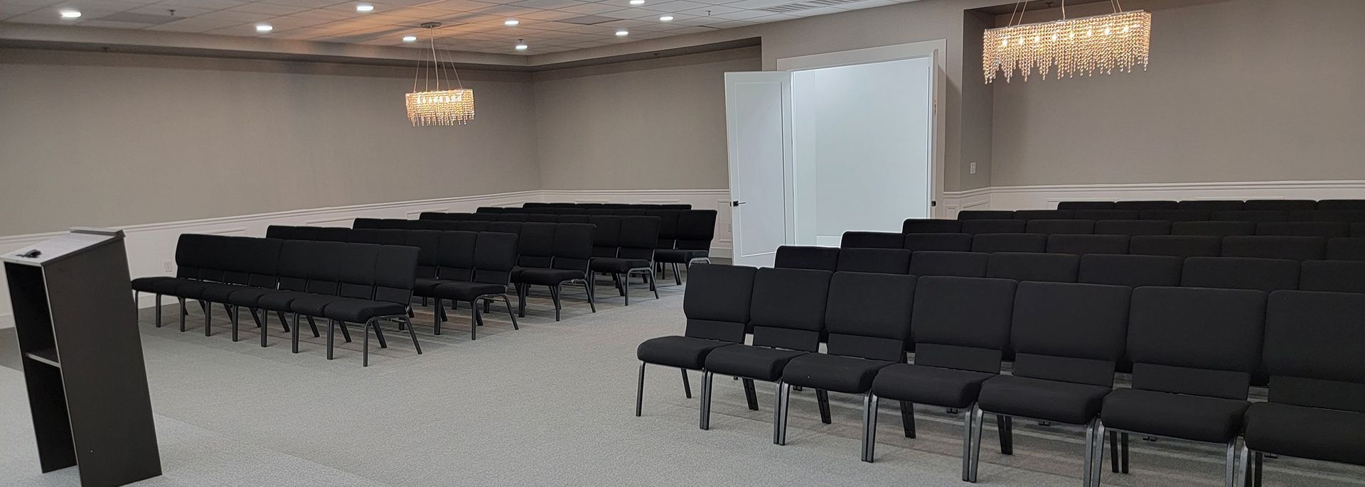 Empty conference room with rows of black chairs, a podium, and a projection screen under warm ceiling lights