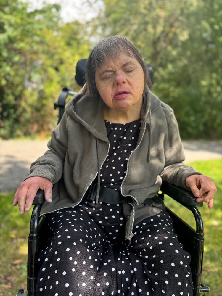 Woman in a wheelchair wearing a gray hoodie and polka dot dress outdoors in front of trees.