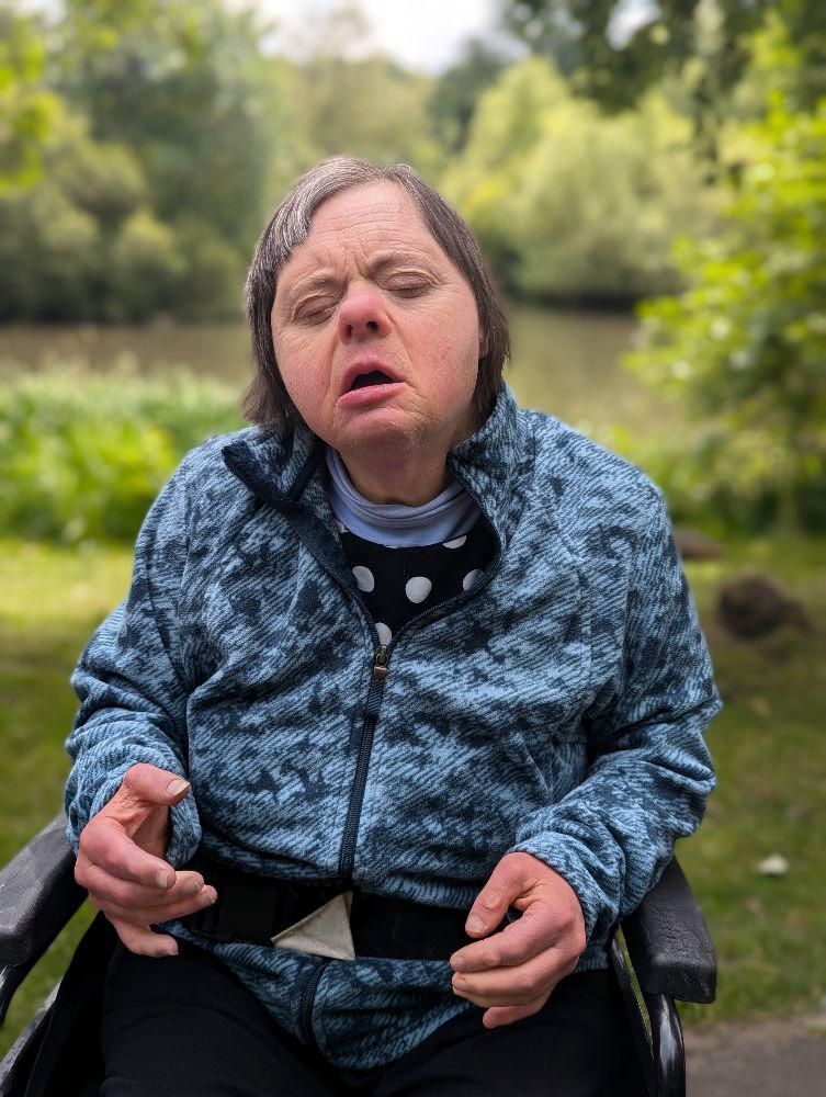 Woman in a wheelchair, wearing a blue jacket, outdoors with trees and a lake in the background.
