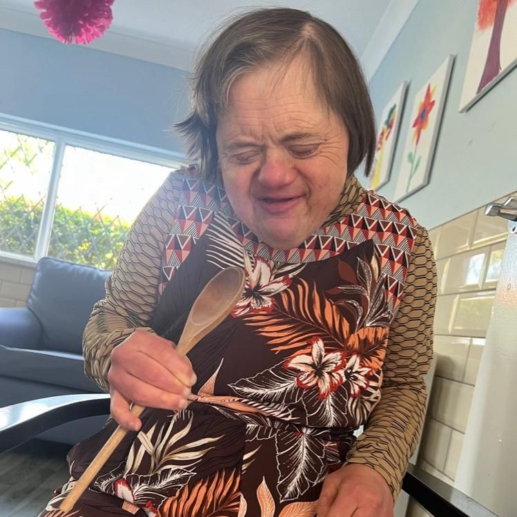 Woman with Down syndrome holds a wooden spoon, wearing an apron, indoors.