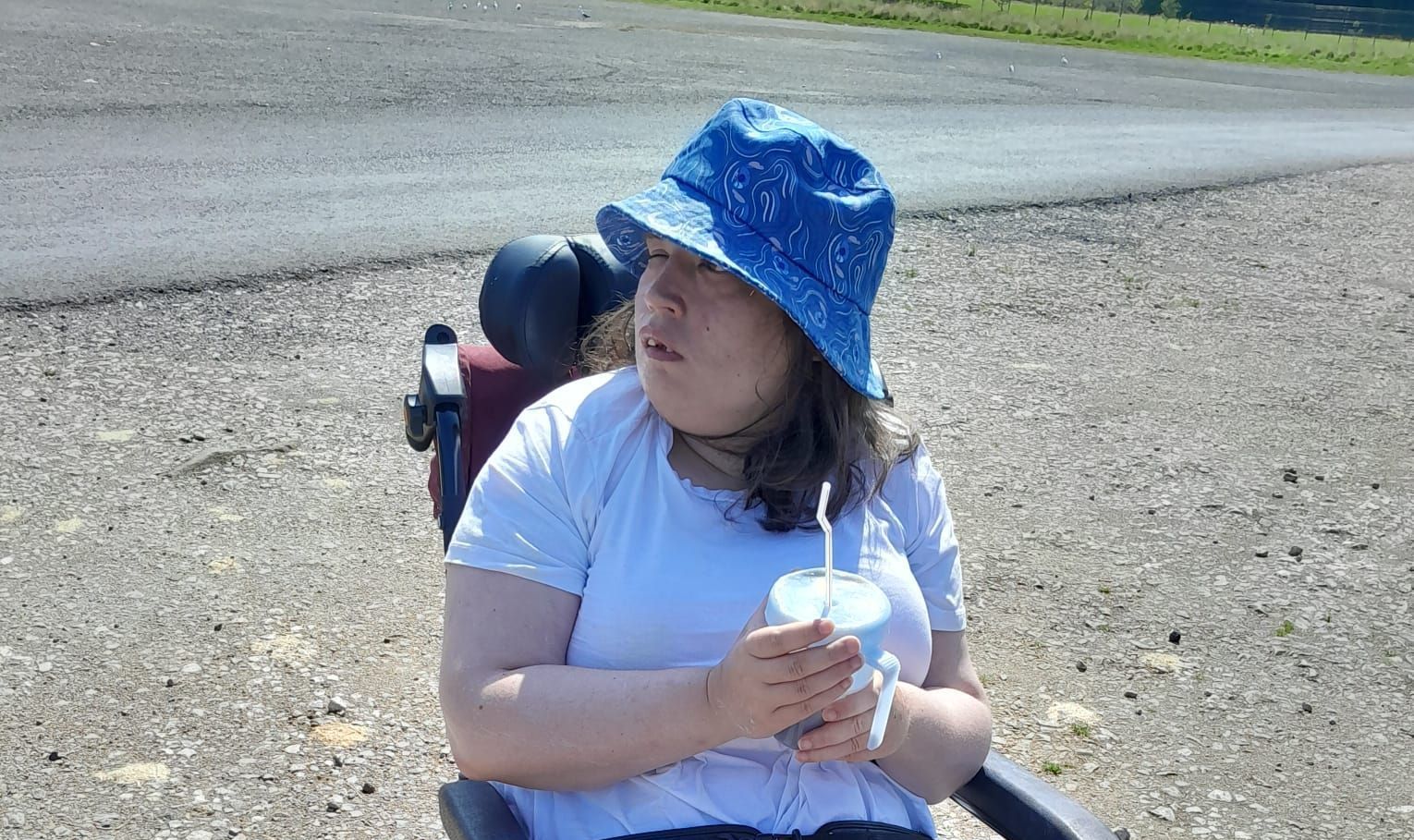 Person in wheelchair wearing a blue hat, holding a drink, looking to the side, outdoors on a sunny day.