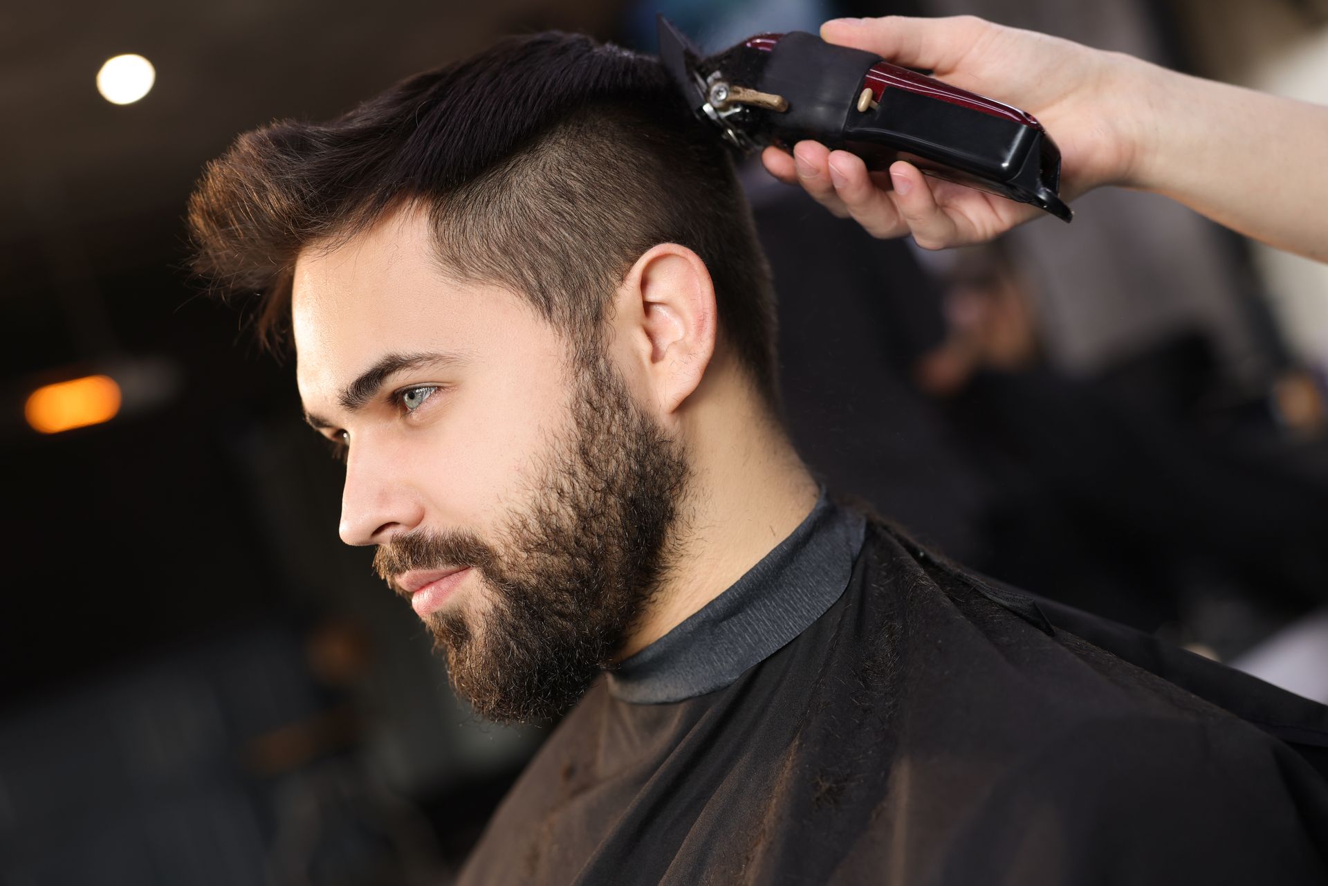 Man getting a haircut, dark hair and beard, clippers in use.