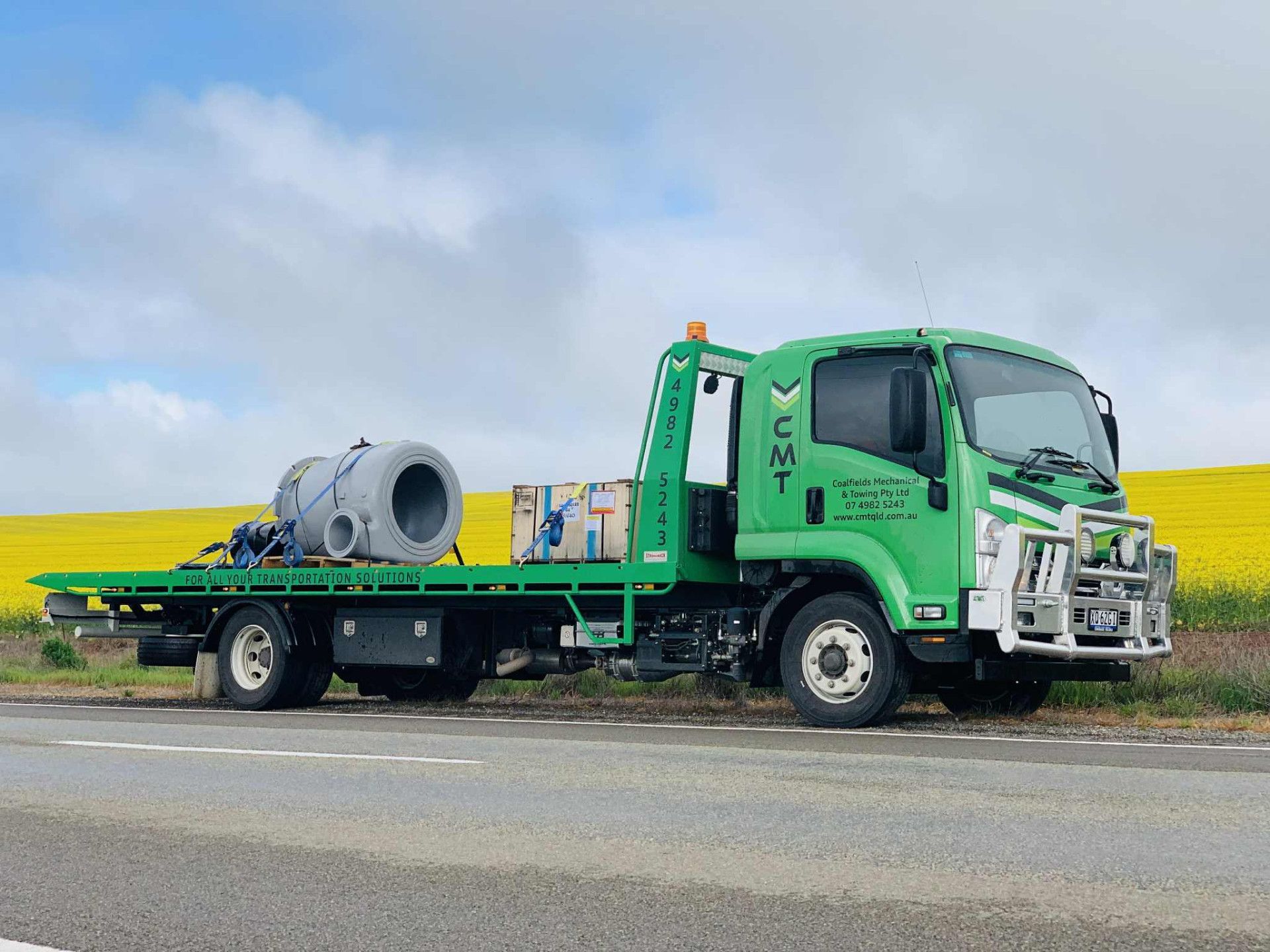 Green CMT truck carrying large gray concrete pipes on a roadside with a yellow flower field background.— Coalfields Mechanical & Towing in Blackwater, QLD