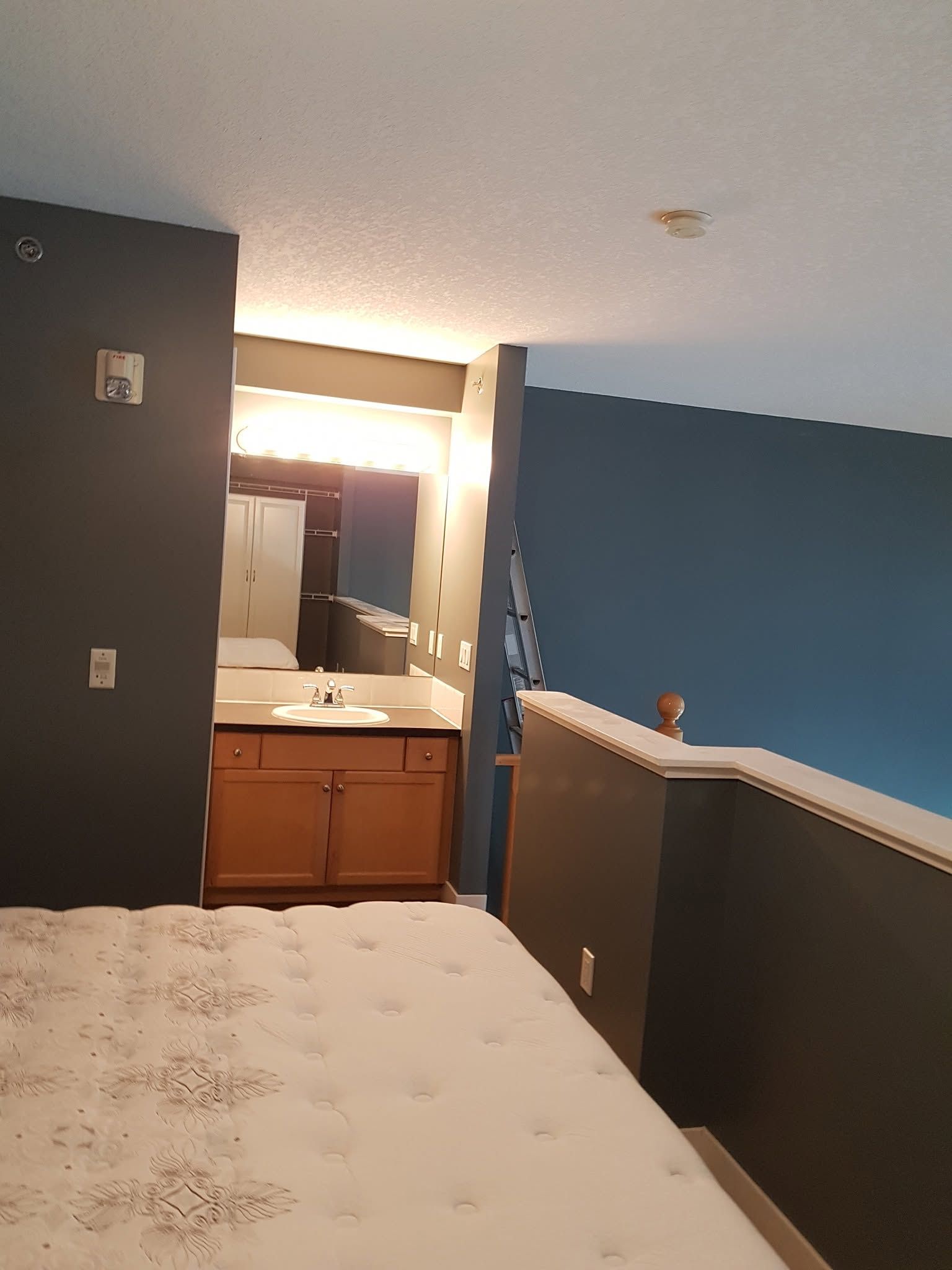 Empty bedroom with blue walls, white bed, and a bathroom vanity visible through the doorway.
