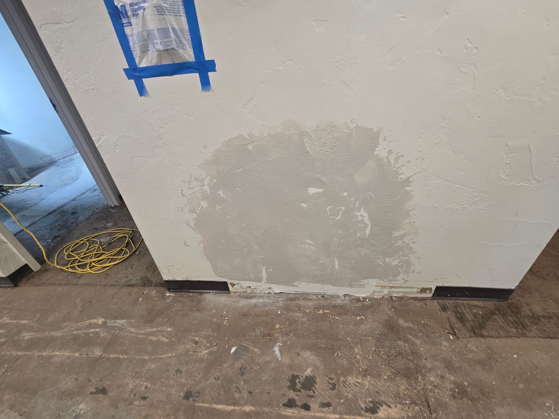 Patchy drywall repair on a white wall near a concrete floor, with blue painter’s tape above.