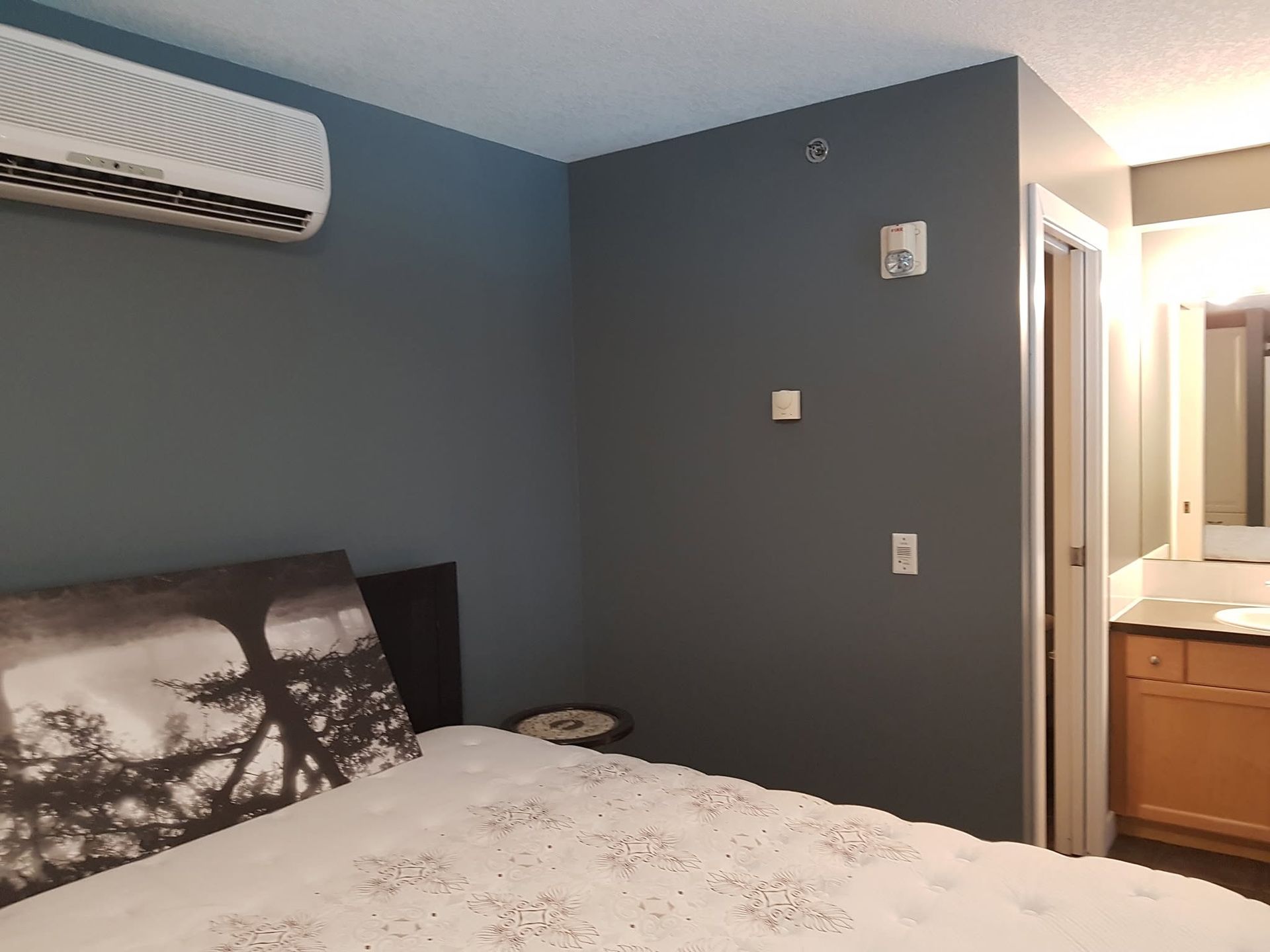 Bedroom with blue-gray walls, bed, wall-mounted AC, and doorway to bathroom vanity
