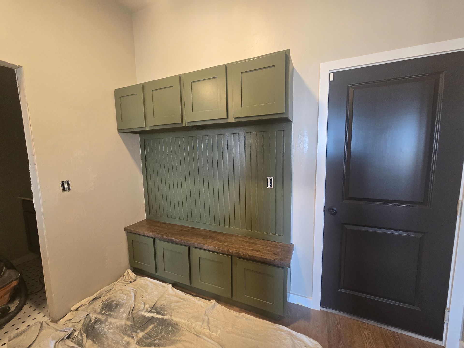Built-in green cabinet and bench with wood top beside a dark door in a room under renovation