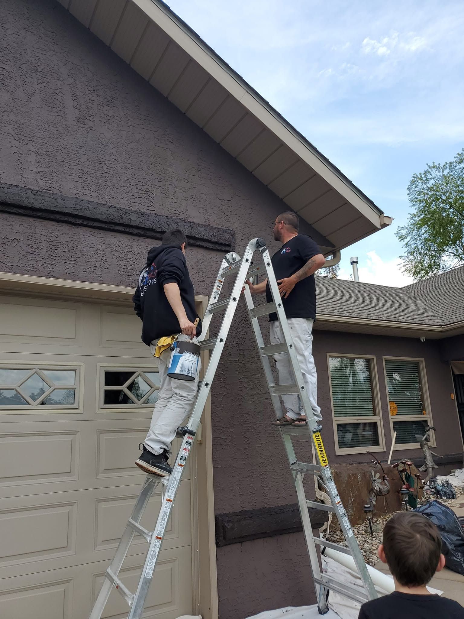 Two workers on ladders repairing the roof and eaves of a house while a child watches below