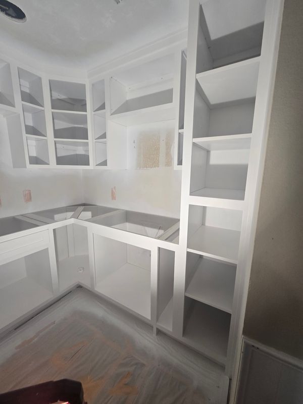 White unfinished kitchen with L-shaped cabinets, open shelving, and gray wood flooring
