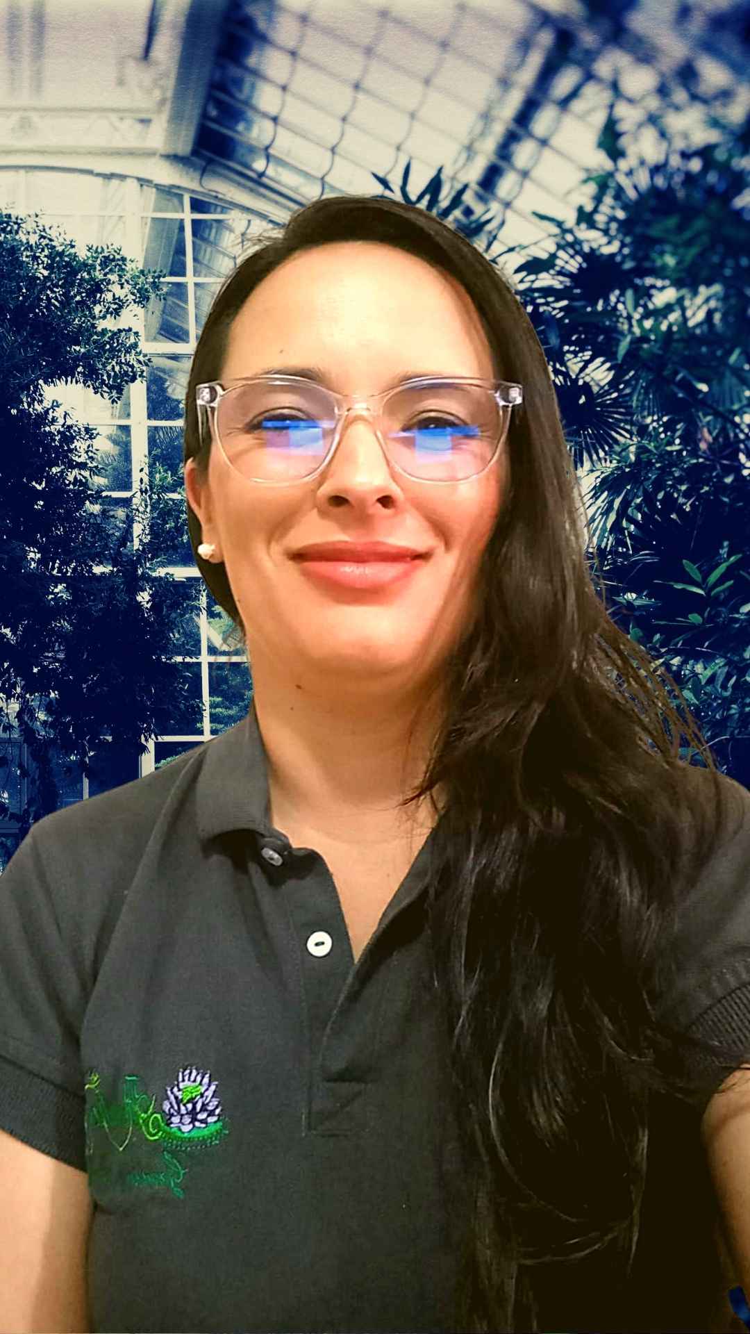 una mujer con gafas y un polo sonríe frente a un invernadero.
