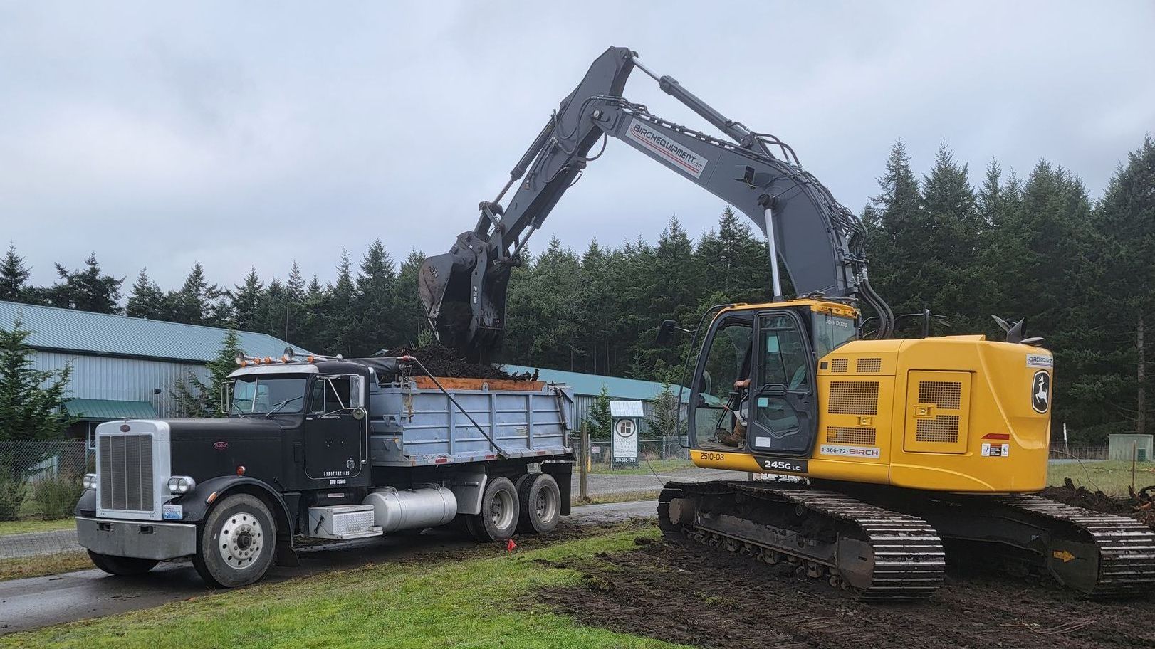 An excavator loads dirt into a black dump truck outdoors under an overcast sky.