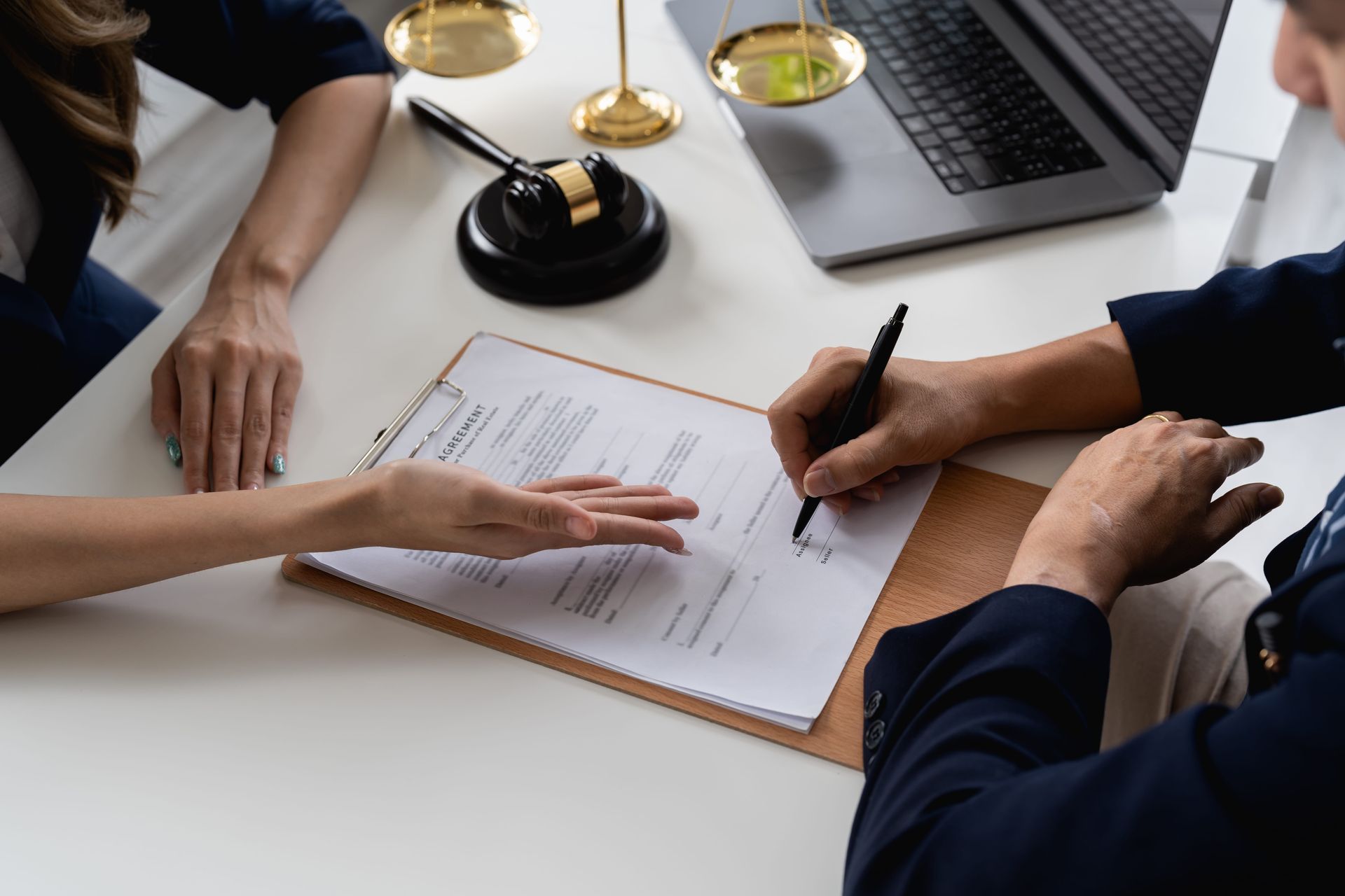 Hands signing a document at a desk with a gavel, laptop, and scales of justice