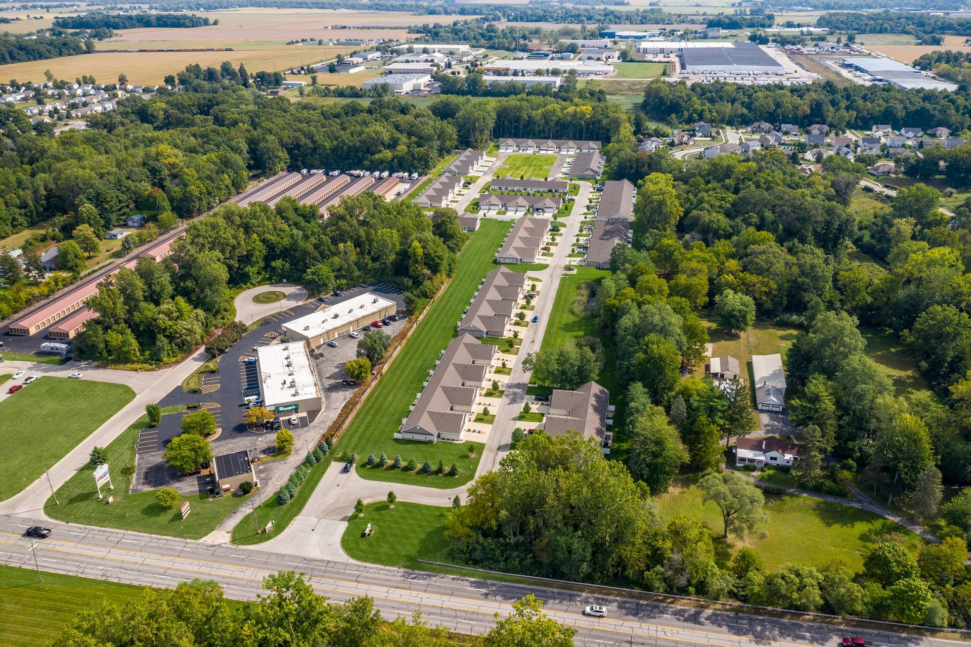 Aerial view of a residential area with townhouses, a commercial building, and green spaces, surrounded by trees and fields.