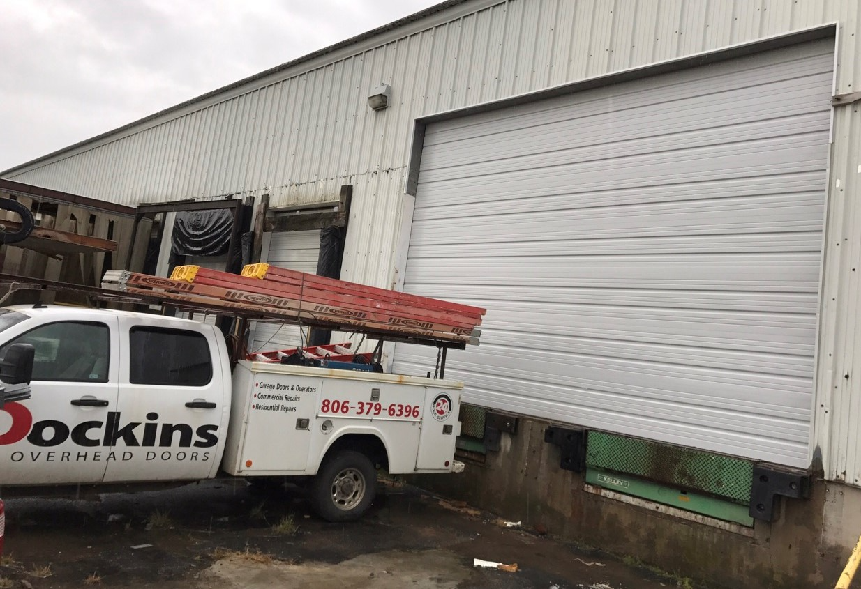 White truck with "Dockins Overhead Doors" parked at a commercial building with a partially open loading dock door.