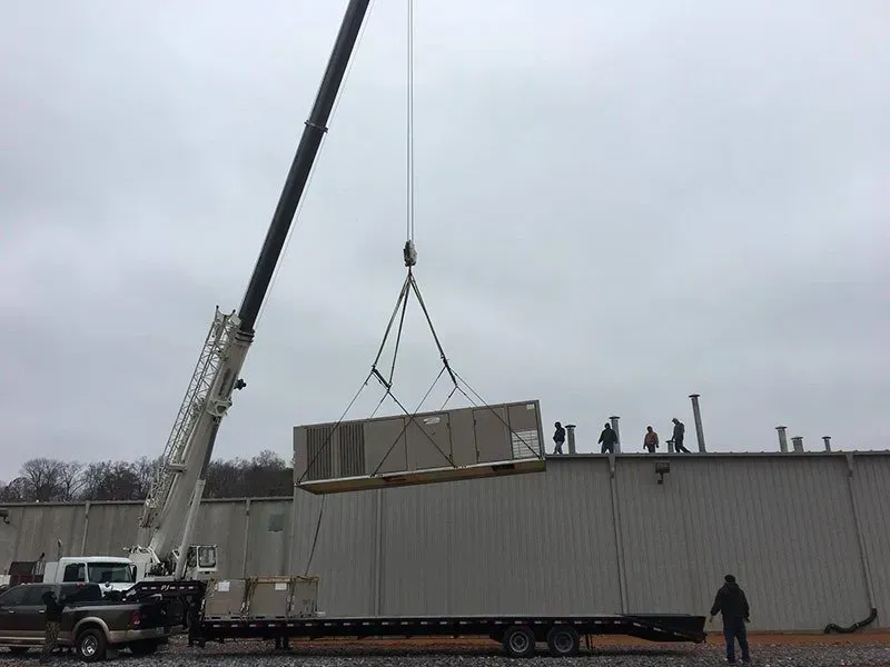 Crane lifting large rectangular unit onto a building roof. Workers observe. Overcast day.