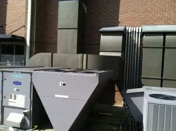 HVAC units and ductwork on a brick building rooftop. Gray metal equipment against brick wall, sunny.