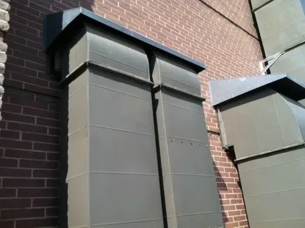Metal air vents attached to a red brick building. Grey rectangular boxes with black tops are secured to the wall.
