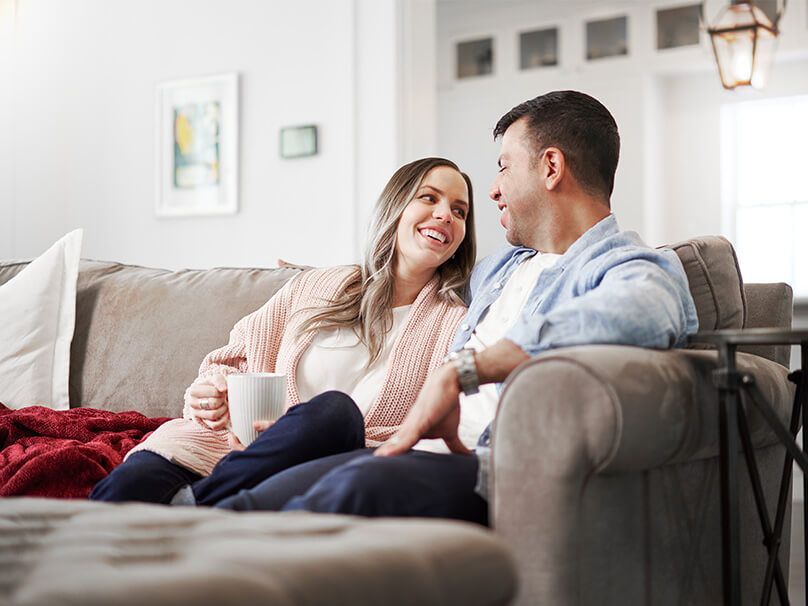 Couple on a sofa, looking at each other and smiling. Woman holds a mug; cozy living room setting.