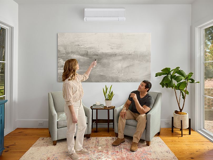 Woman adjusting an AC unit's settings with a remote. Man watches from a chair in a bright living room.
