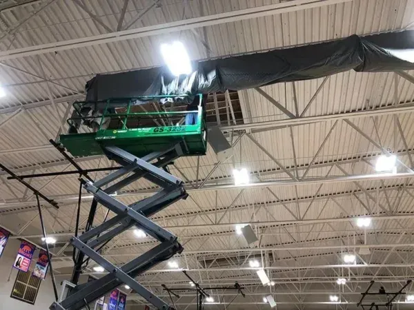 Green lift holds a person working near the gymnasium ceiling, next to a long dark covering.