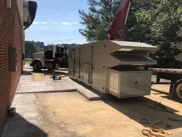 A large HVAC unit being lifted by a crane near a brick building and a truck.