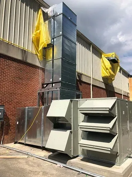 Exterior HVAC system with metal ductwork rising above a brick wall and a concrete base. Yellow protective covers are attached.