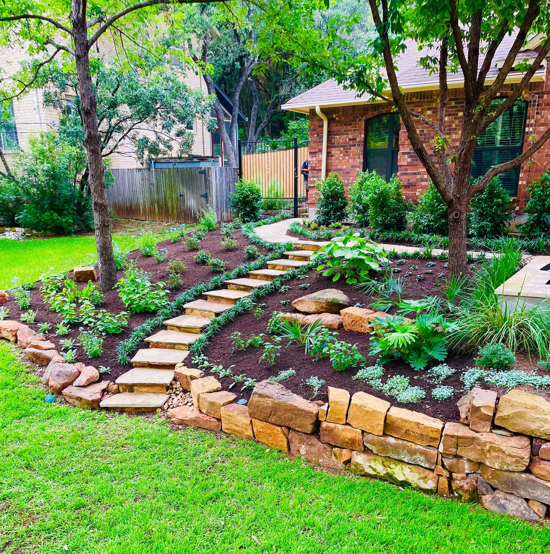 Stone garden path with steps, terraced flower beds, and a brick house under leafy trees