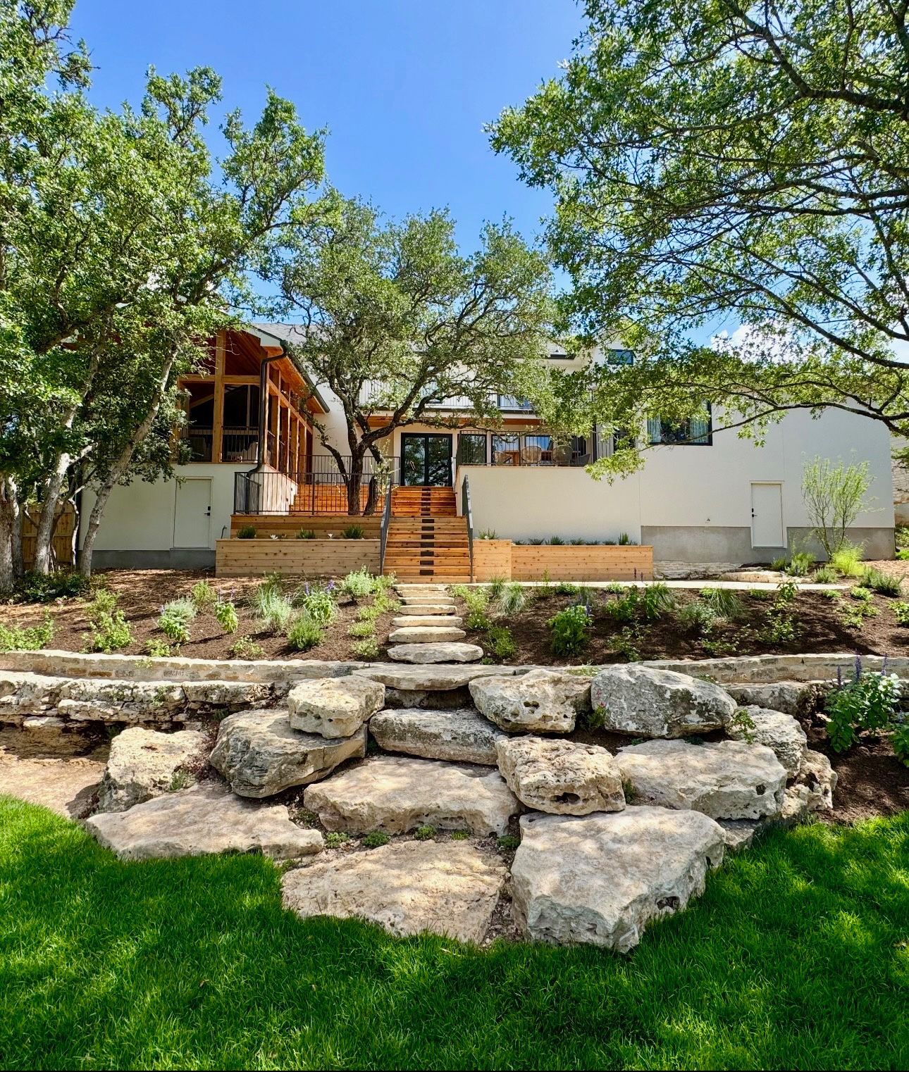 Modern house with wood deck and stairs, framed by trees and a rocky landscaped foreground under a blue sky
